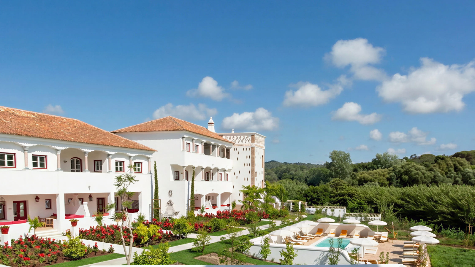 Lush garden and pool complex at the Vermelho Melides white villa under a blue sky with clouds. Forested background creates a serene mood.