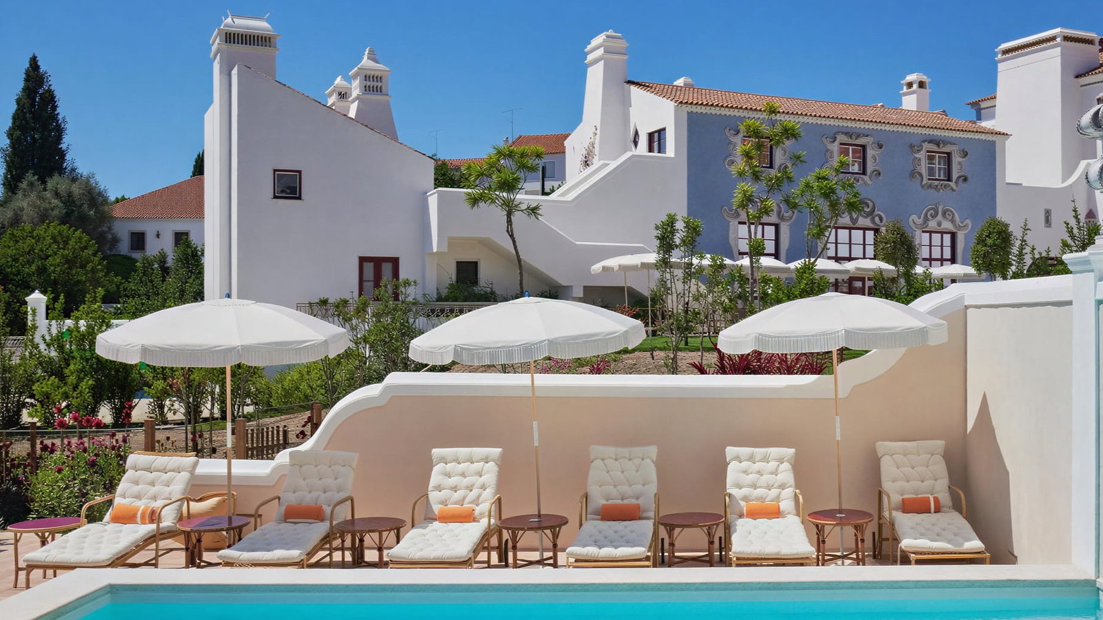 Vermelho Melides pool with six lounge chairs and umbrellas, set against white and blue buildings, lush gardens, and a clear blue sky. Relaxing mood.