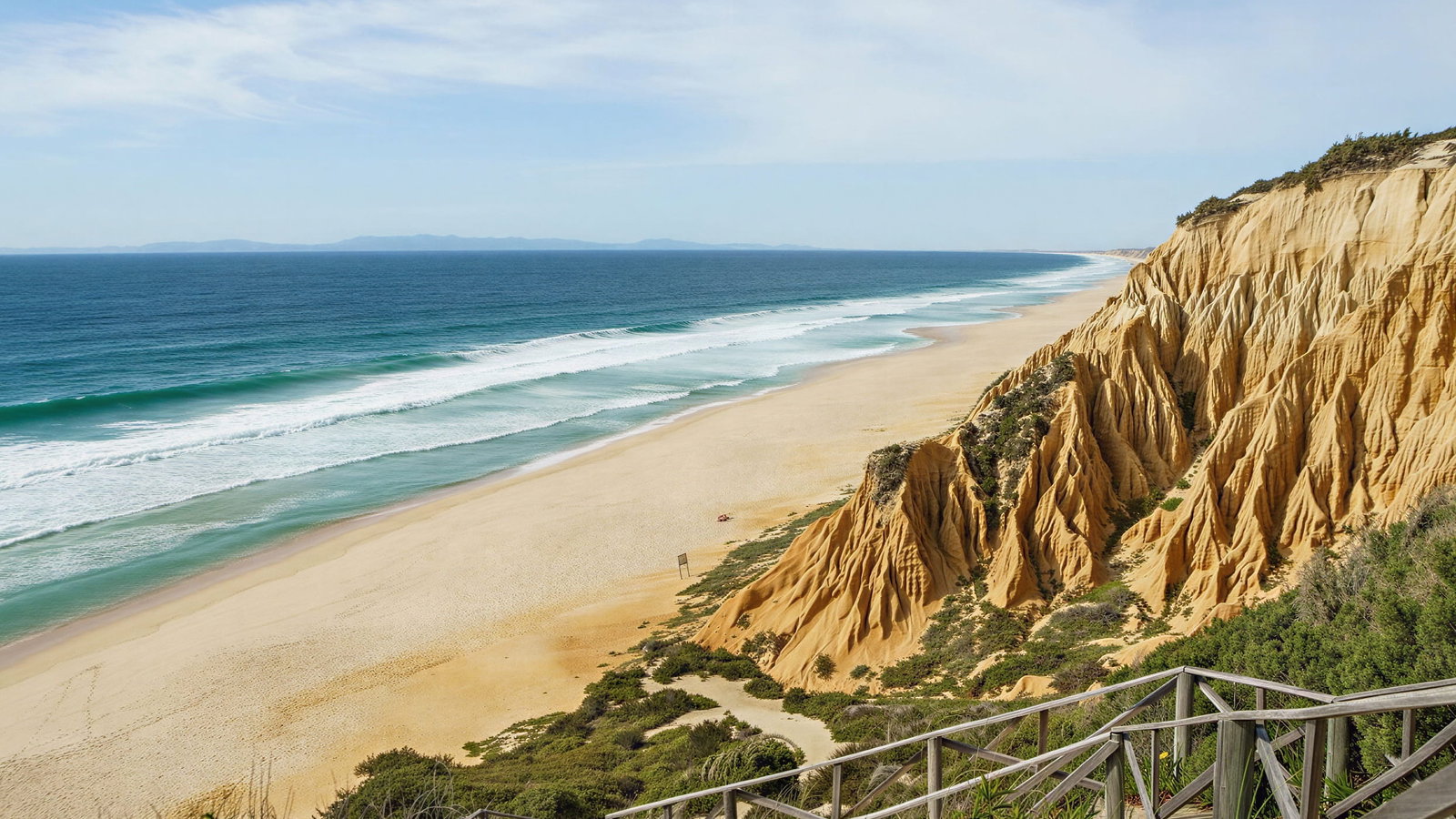 Praia da Gale Beach near Vermelho Melides. Stunning coastal view of sandy cliffs and a wooden walkway leading to a vast beach with turquoise waves under a clear blue sky.