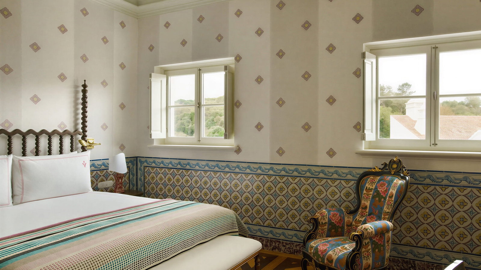 Elegant Junior Suite bedroom at Vermelho Melides with patterned wallpaper, ornate bed, colorful armchair, and two windows overlooking greenery. Calm, inviting ambiance.