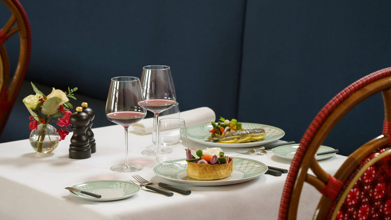 Elegant Xtian restaurant table with white cloth at Vermelho Melides, two plates of gourmet food, two glasses of red wine, a vase with flowers, and red chairs.