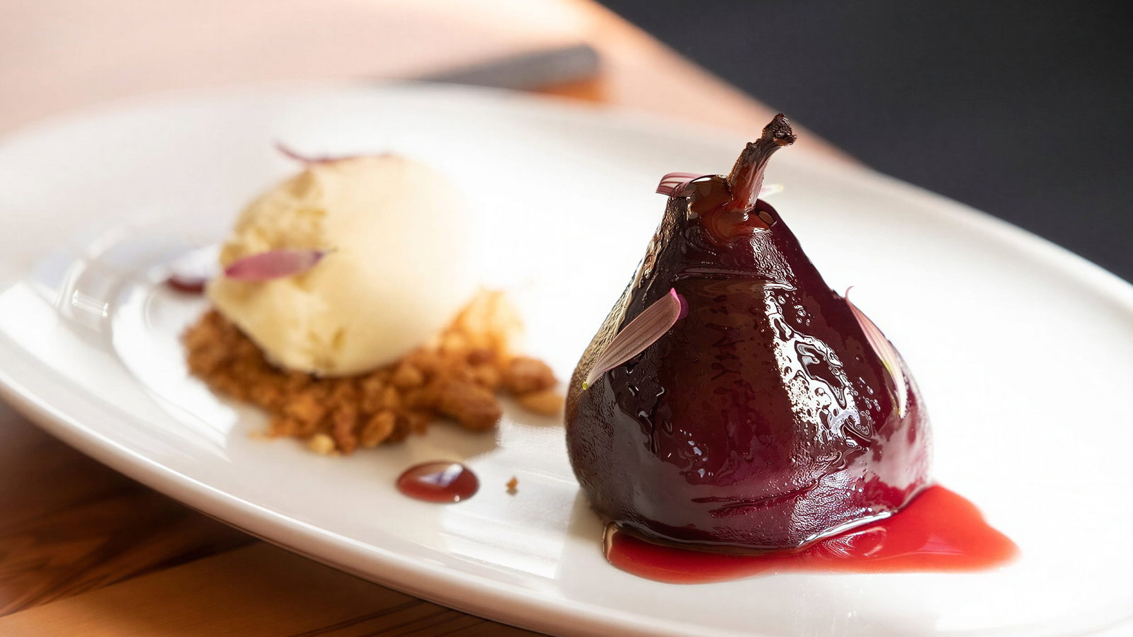 Dessert from Restaurante Xtian at Vermelho Melides. Poached pear in dark glaze and vanilla ice cream with crumbles on white plate. Wooden table setting, warm lighting. Elegant dessert presentation.