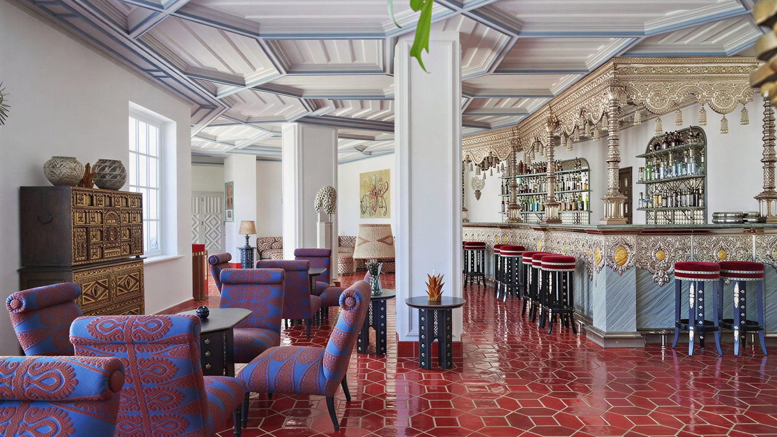 Elegant bar interior at Vermelho Melides with ornate decor, red hexagonal floor tiles, and blue patterned chairs. A vintage cabinet and vibrant furnishings complete the scene.