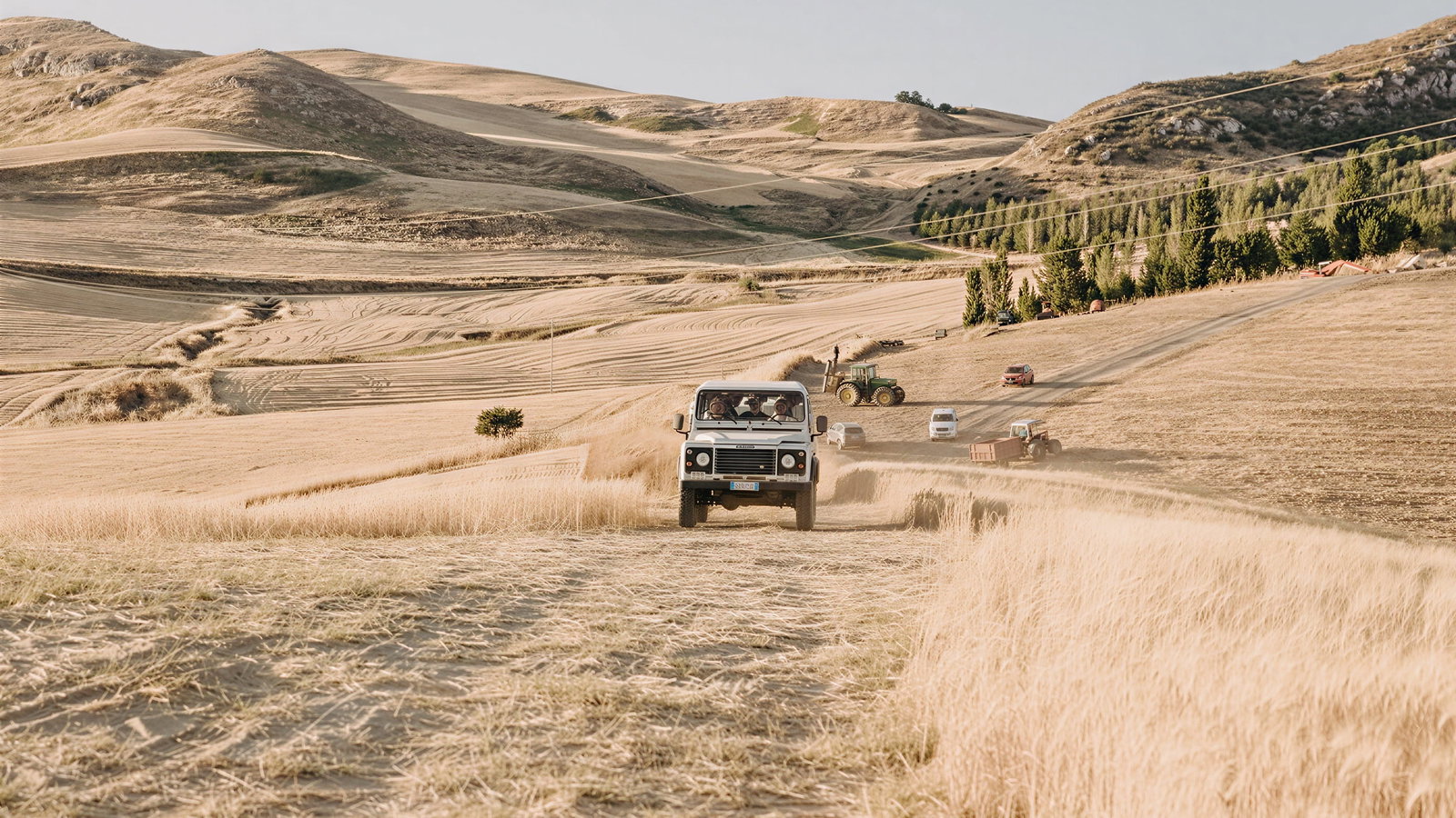 Jeep tour Masseria Susafa, driving through golden fields with dust trail. Hills and tractors in background. Clear sky, rural setting, conveying adventure.