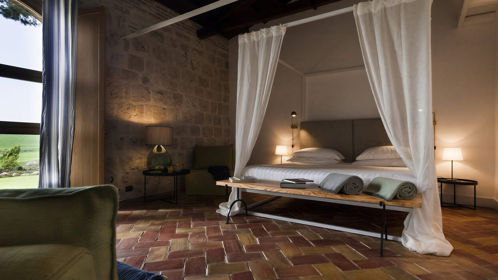 Classic suite bedroom at Masseria Susafa with a canopy bed, rolled towels, and soft lighting. Brick floor, stone wall, curtain, and window view of greenery.
