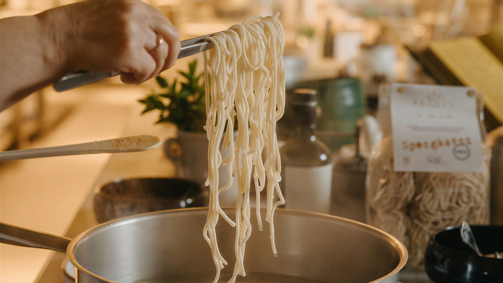 Fresh pasta workshop at Masseria Susafa. A hand holding tongs lifts spaghetti from boiling pot; a pan of sauce in foreground. Kitchen background with warm lighting, pasta packet visible.