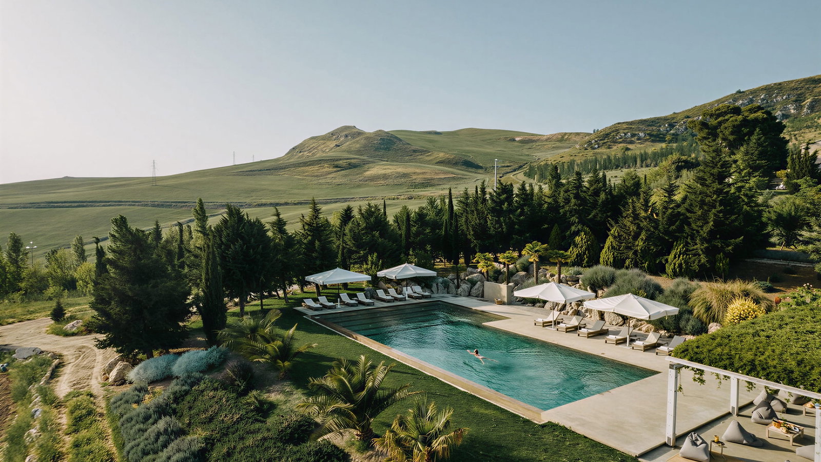 The pool at Masseria Susafa, an oasis with loungers and umbrellas, surrounded by lush greenery and hills. A person swims in clear water under a blue sky.