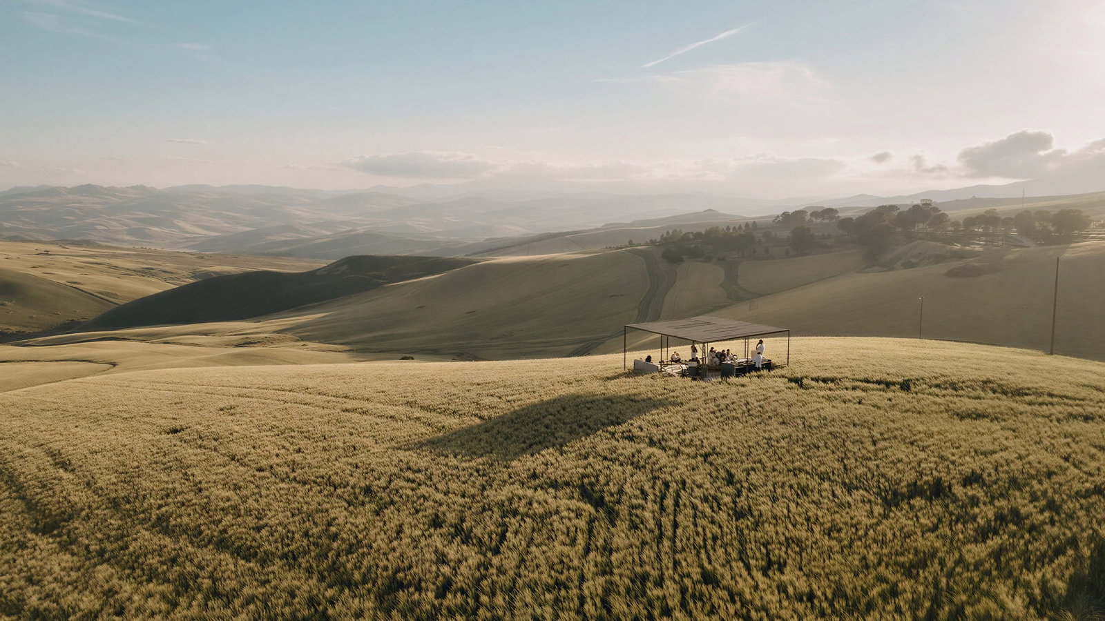 Picnic in the fields at Masseria Susafa under a canopy on a rolling hillside, surrounded by golden fields and distant mountains, with a clear blue sky above.