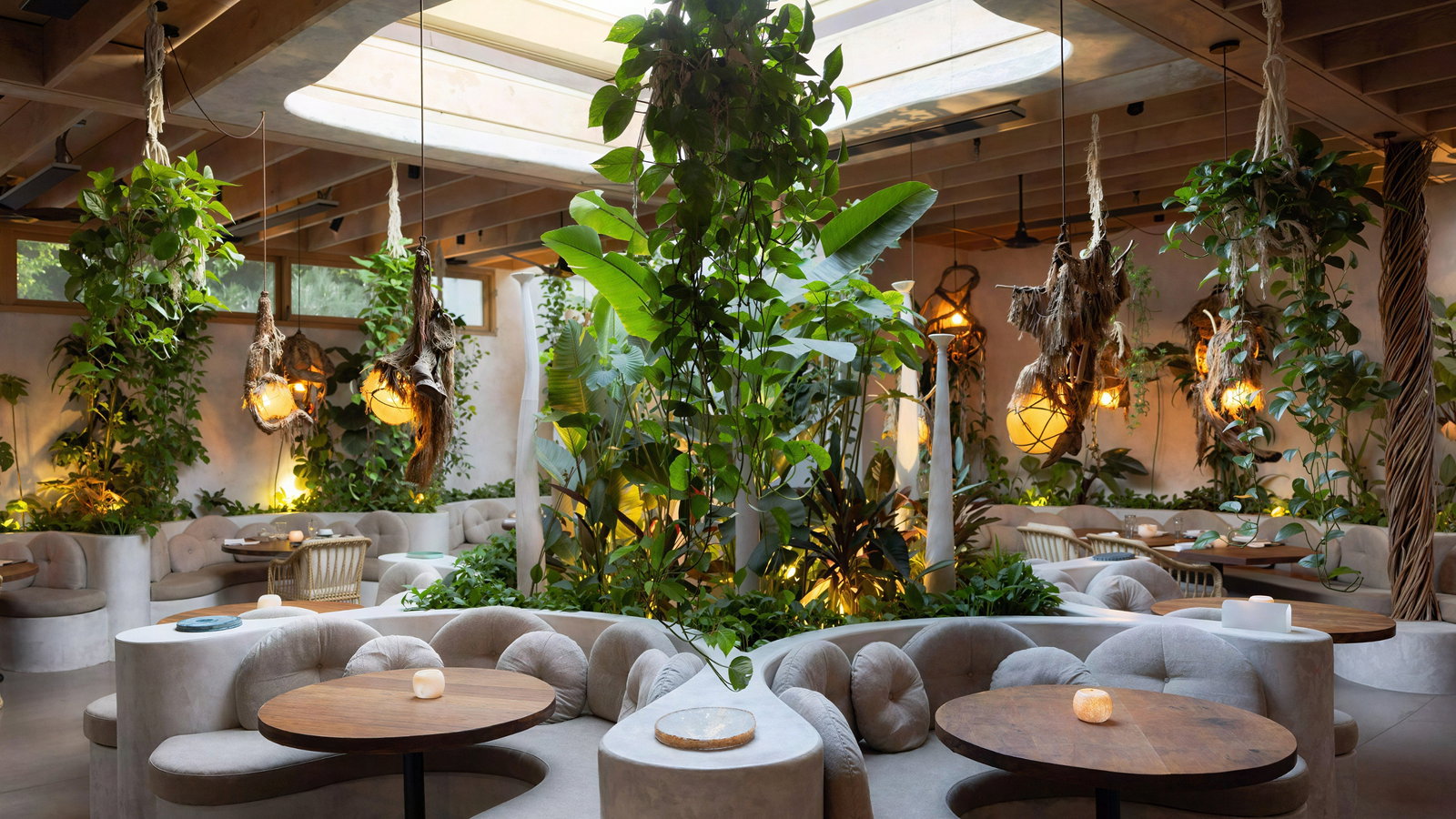 Indoor restaurant with lush greenery, round tables, and cushioned seating. Soft lighting creates a cozy, inviting atmosphere.