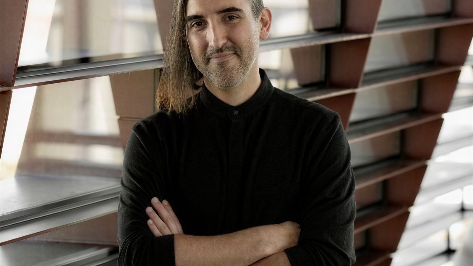 A person stands with arms crossed, smiling slightly. They wear a black shirt and have long hair. Background features geometric window designs.