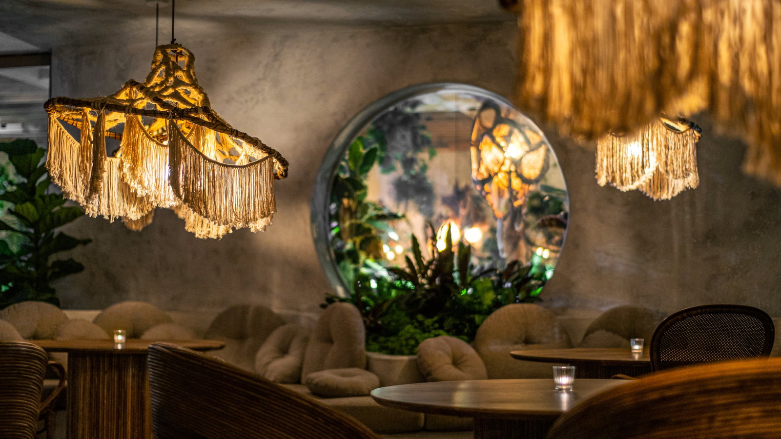 Cozy café with beige fringed lamps, round wooden tables, and plush seating. A circular mirror reflects plants and warm lighting.