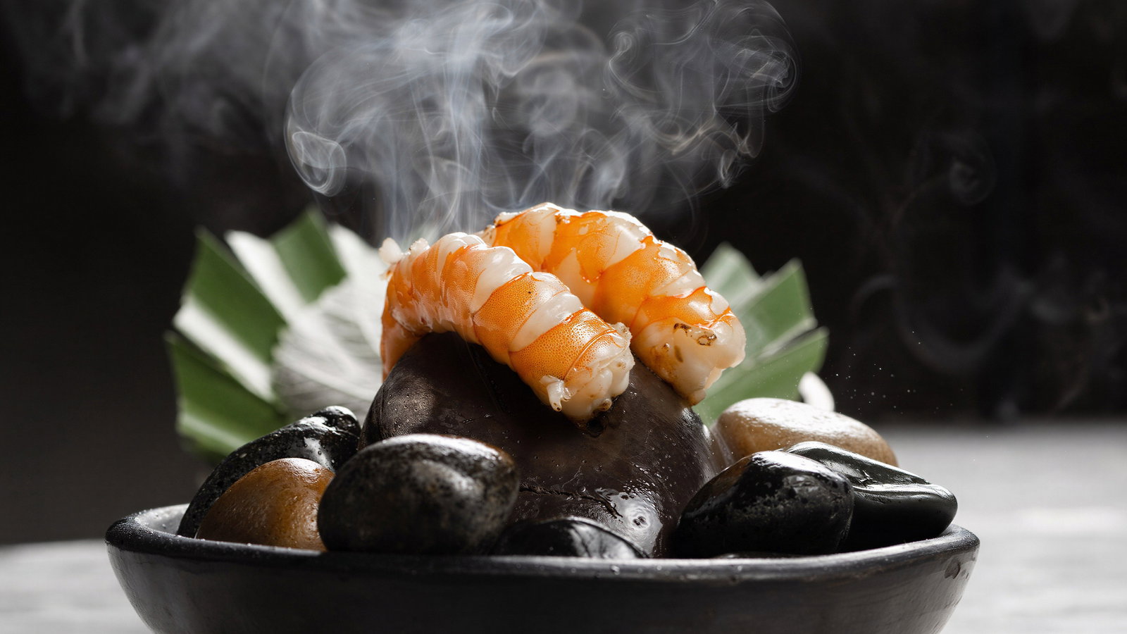 Steaming shrimp on black stones in a dark dish, with steam rising, set against a blurred green leaf and dark background.