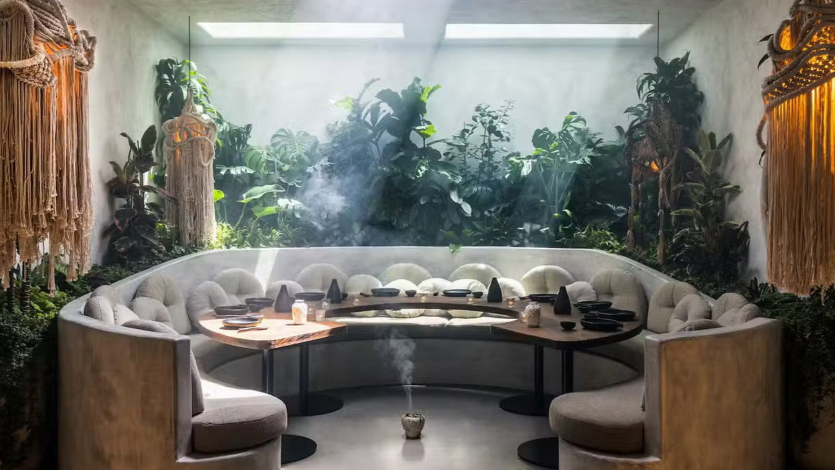 Cozy seating area with round sofa, wooden table, and smoky incense. Lush plants and woven decor under a skylight create a serene mood.
