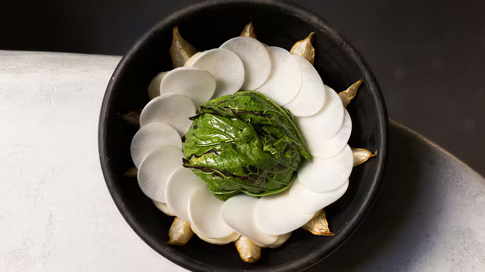 A black bowl holds a decorative arrangement of white radish slices around a center of green leafy vegetables, on a textured white surface.