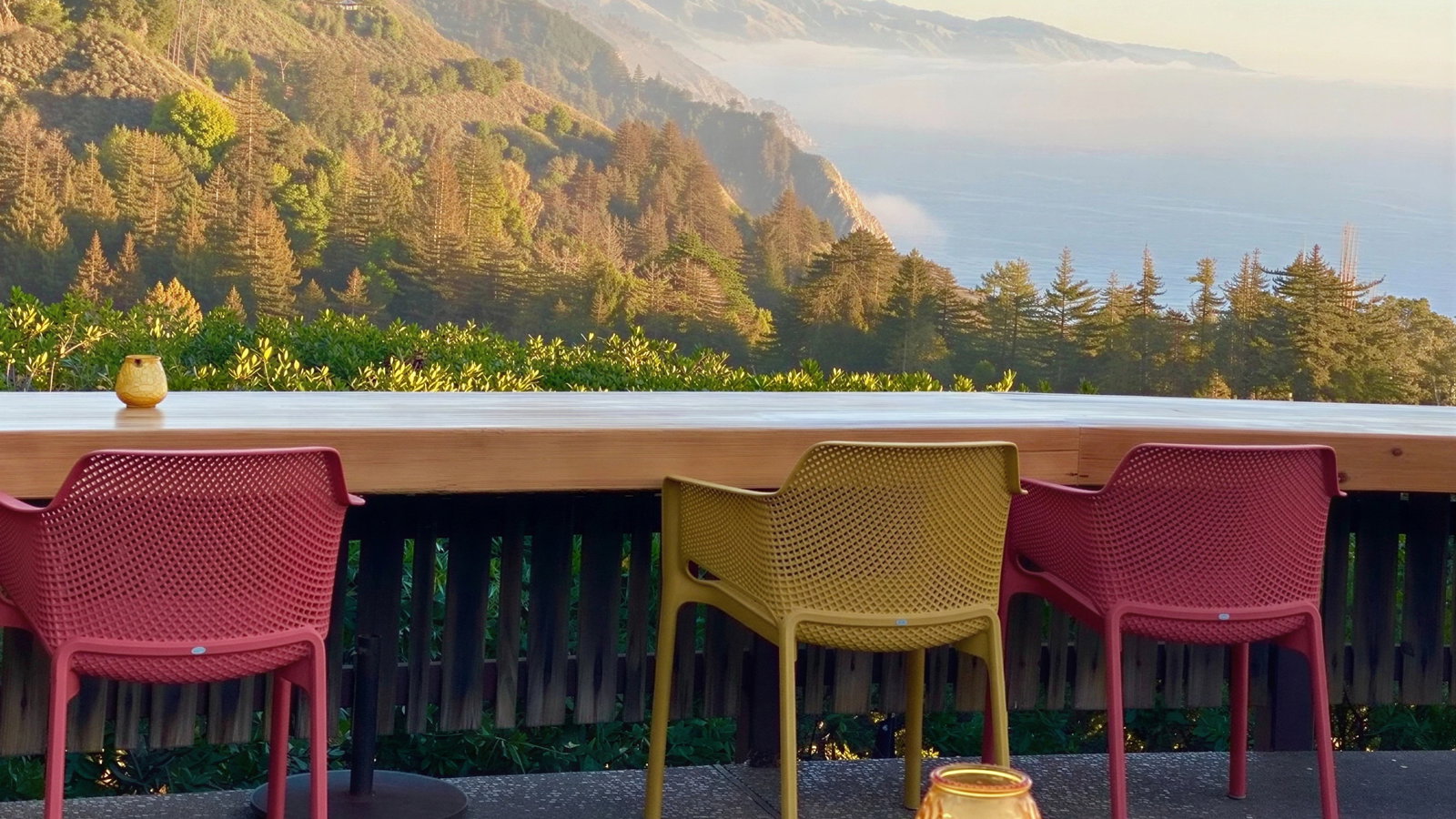 Colorful chairs and candles sit on a patio overlooking a misty coastal forest and mountain range under a clear sky.