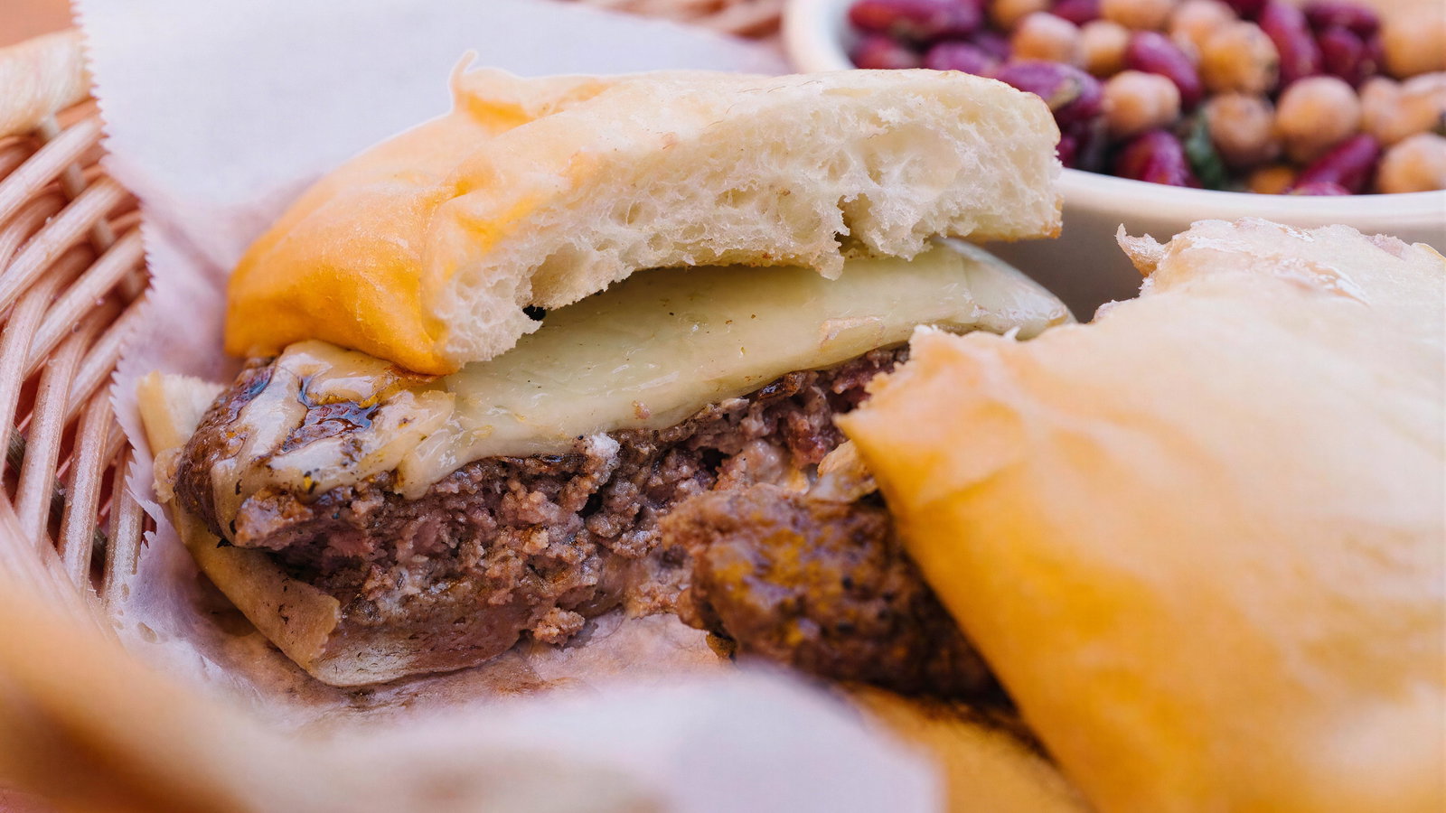 Burger with melted cheese in a basket, next to a bowl of chickpeas and kidney beans. Bright, appetizing setting with a casual vibe.