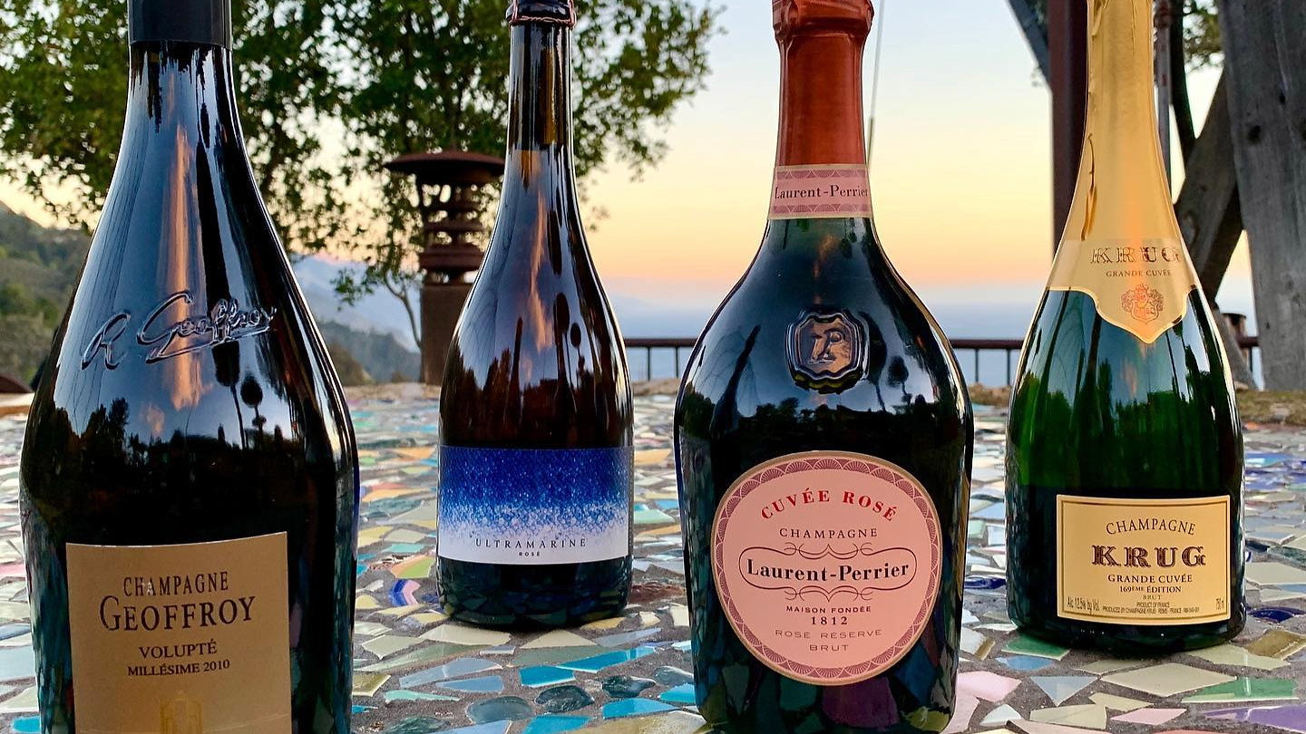 Four champagne bottles on a colorful mosaic table with a scenic sunset view and trees in the background. Text includes Geoffroy and Krug.