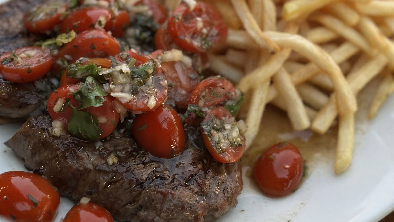 Steak topped with cherry tomatoes and herbs on a white plate, accompanied by golden fries. The vibrant colors suggest a savory meal.