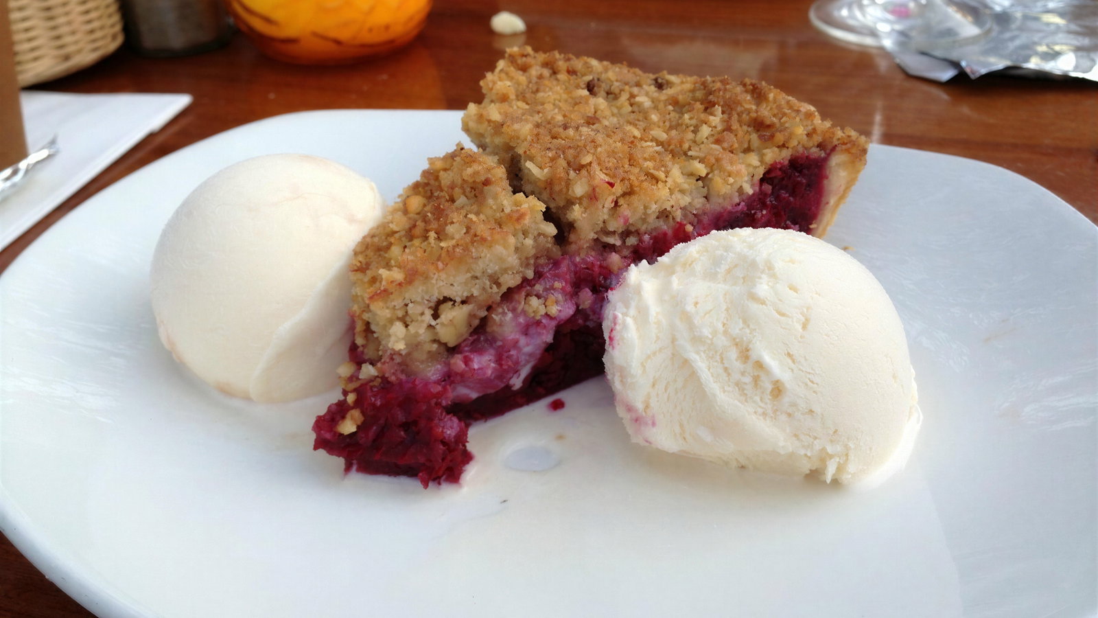 A slice of berry crumble with two scoops of vanilla ice cream on a white plate, set on a wooden table with a yellow candle in the background.