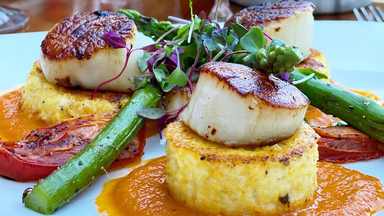 Seared scallops on risotto cakes with asparagus, tomato, and greens, set on orange sauce. Vibrant colors on a white plate.