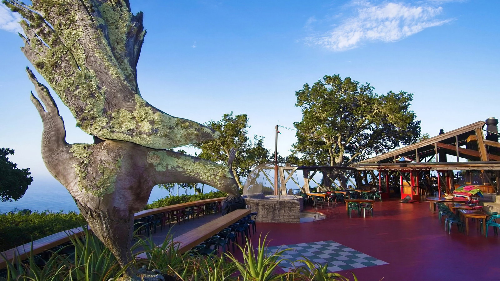 Outdoor patio with colorful tables and chairs, large bird sculpture, and ocean view. Sunny, clear sky with a few clouds.