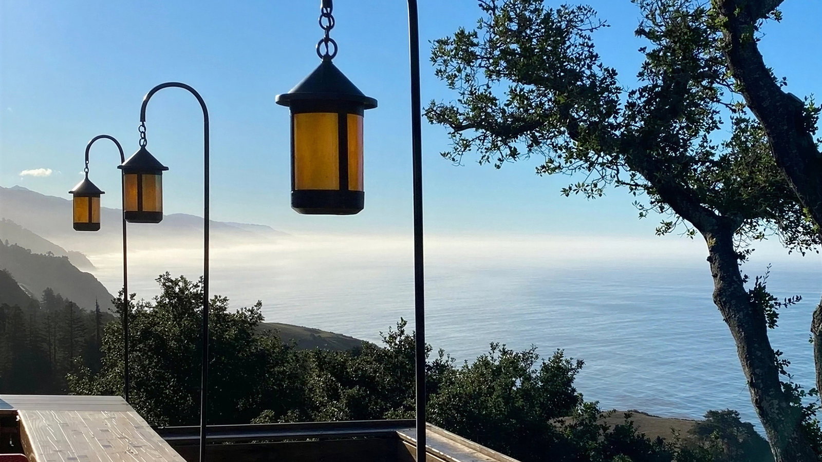 Vintage lanterns along a wooden deck overlook a misty ocean and mountainous landscape, with trees silhouetted against a clear blue sky.
