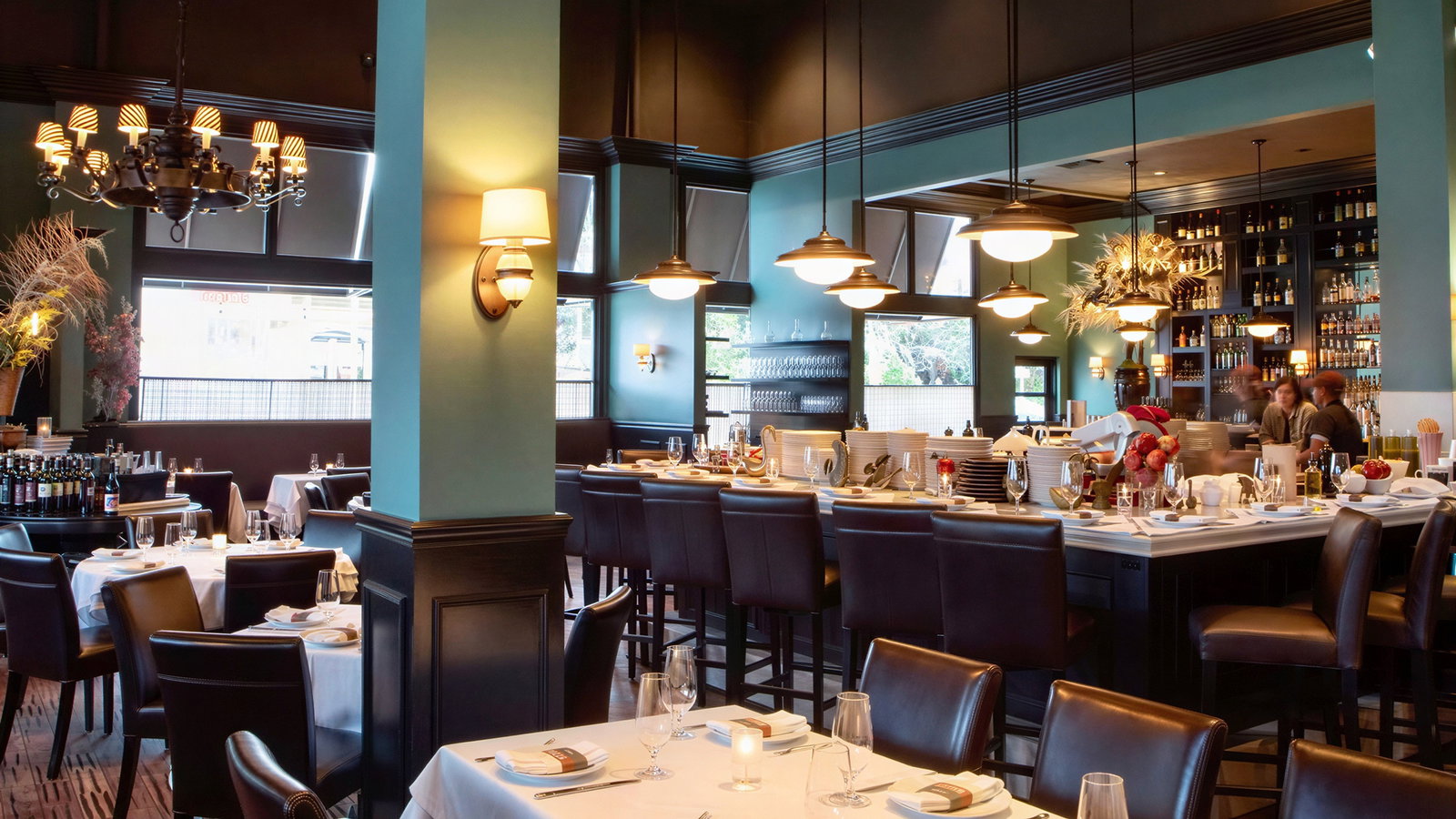 Elegant restaurant interior with teal walls, dark wood furniture, and chandeliers. Tables are set with white linens and glassware.