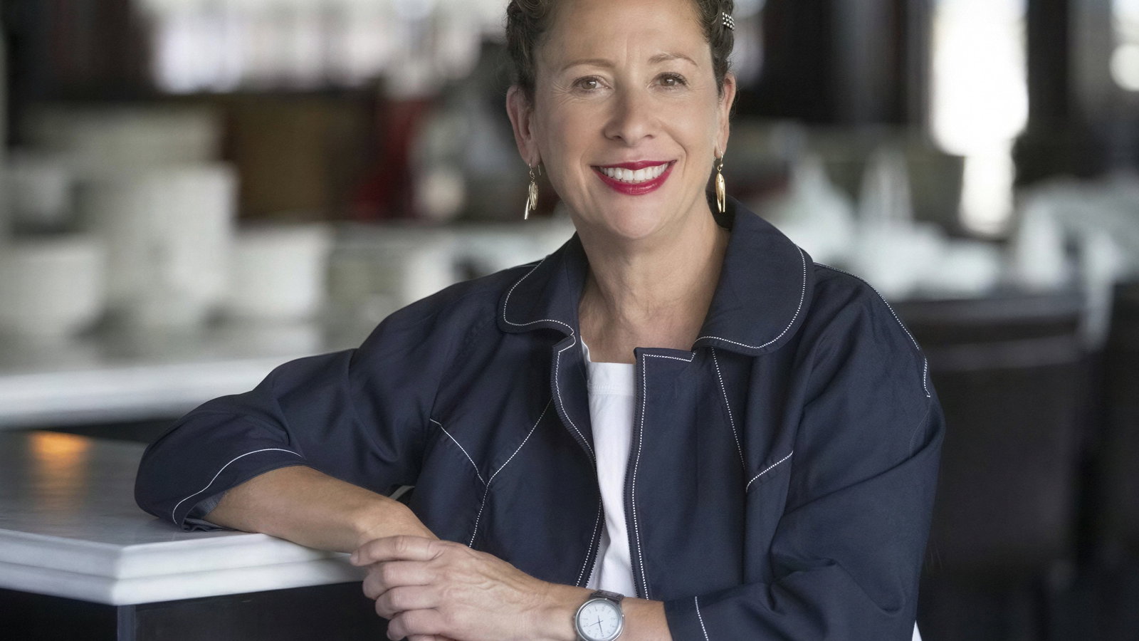 Smiling woman in a navy jacket leans on a white table in a softly-lit room. She wears a watch and hoop earrings, creating a welcoming mood.