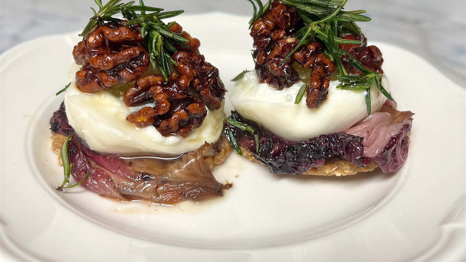 Two appetizers on a white plate with cheese, candied nuts, and fresh rosemary atop radicchio. Set against a marble background.