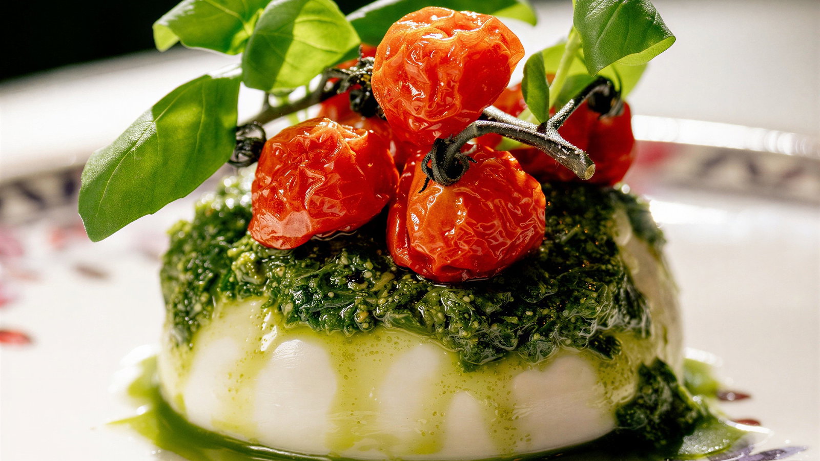 Pesto-covered burrata topped with roasted cherry tomatoes and basil leaves on a floral-patterned plate, creating a fresh, vibrant presentation.