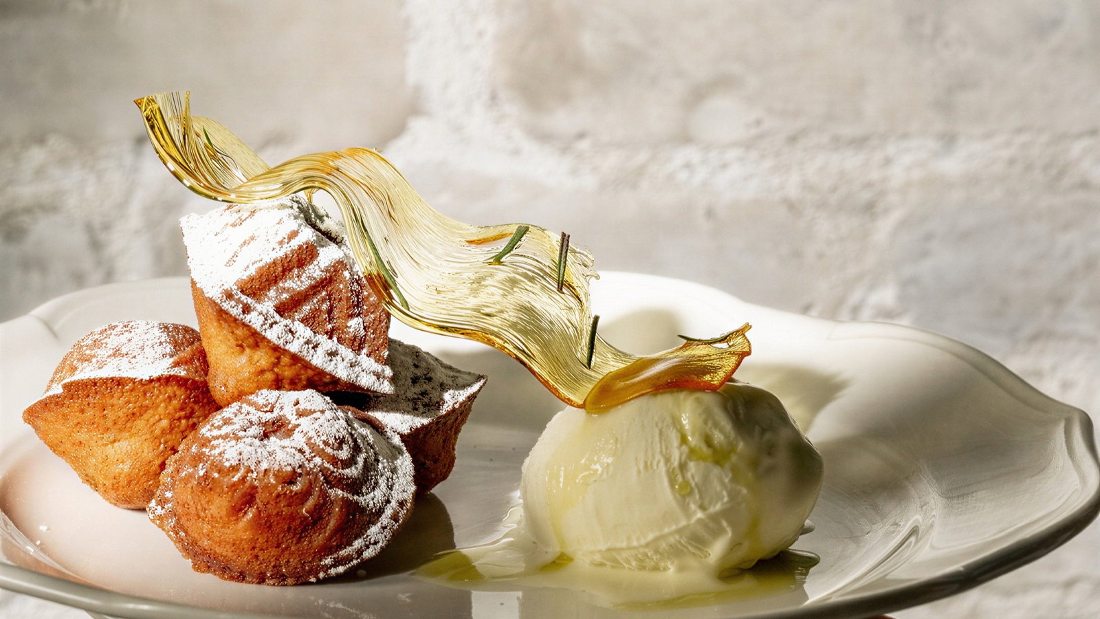 Madeleines dusted with powdered sugar beside vanilla ice cream on a white plate, garnished with golden decoration. Bright, textured background.