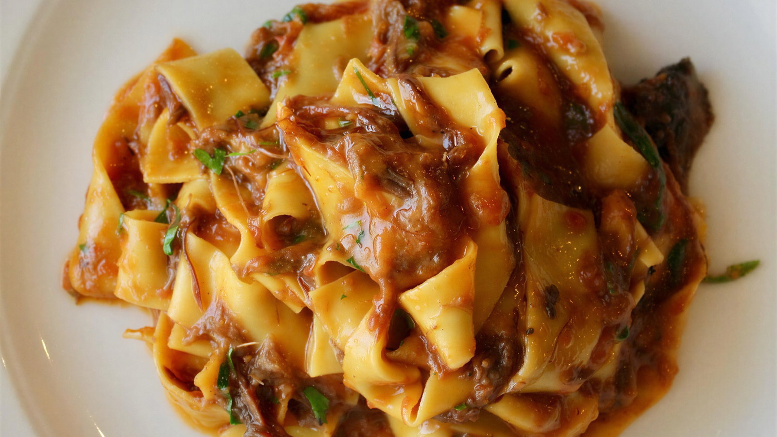 Close-up of pappardelle pasta with rich meat sauce and herbs on a white plate. The dish has a hearty, appetizing appearance.
