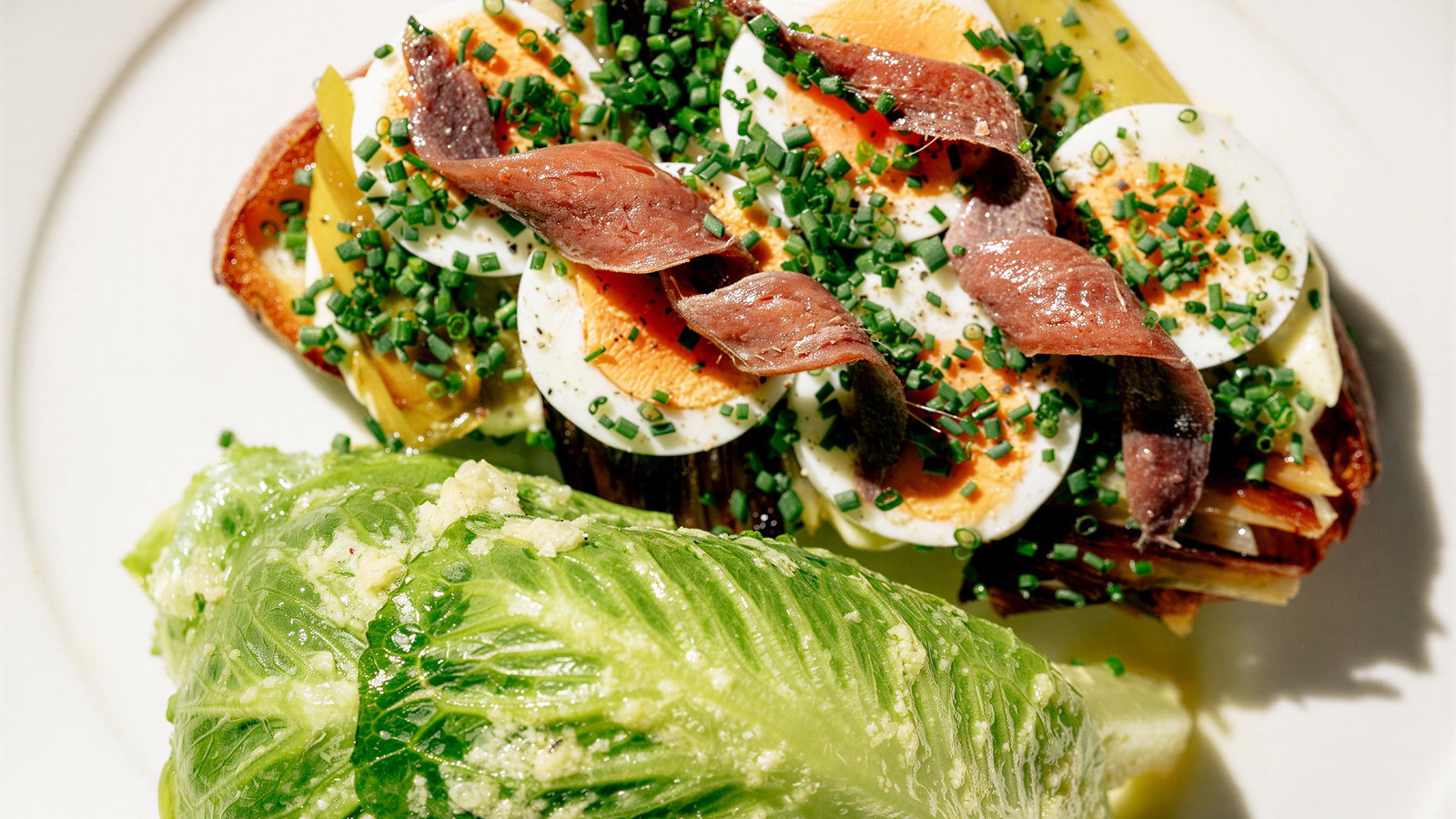 Toast topped with sliced eggs, anchovies, chives, and roasted vegetables beside a leaf of fresh romaine lettuce on a white plate.