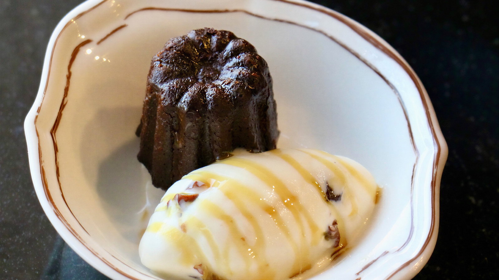 A dessert plate with a chocolate canelé and vanilla ice cream drizzled in honey. Set against a dark background, evoking an indulgent mood.