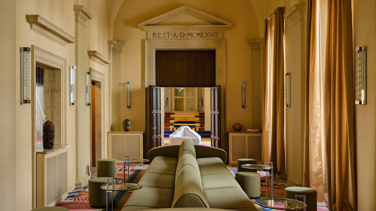 Palazzo Talìa lobby room with arches, beige walls, and brown curtains. Modern green sofas and colorful patterned carpet. Text above door: "REST·A·D·MCMLXVII".