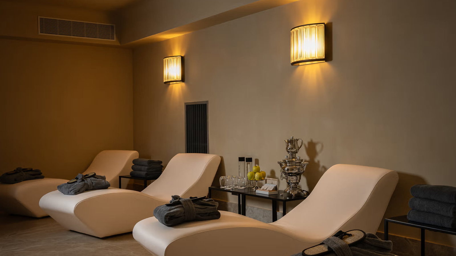 Four cream lounge chairs in the relaxing area of Palazzo Talìa spa with dark robes and towels in a dimly lit spa room. Yellow wall lights create a serene ambiance.