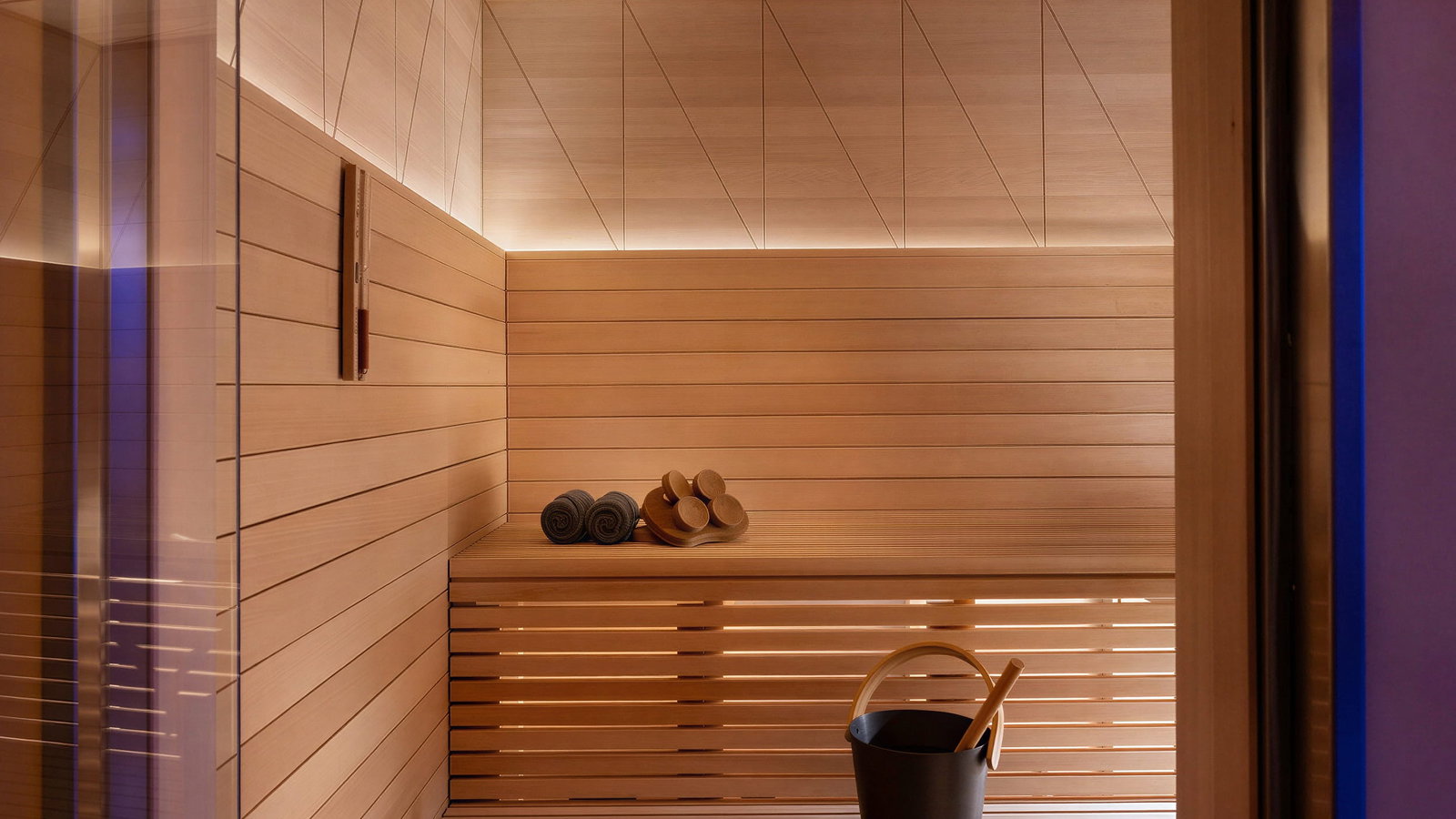 Wooden sauna interior at Palazzo Talìa with two rolled towels, sauna rocks, and a black bucket on wooden benches. Warm lighting creates a cozy ambiance.
