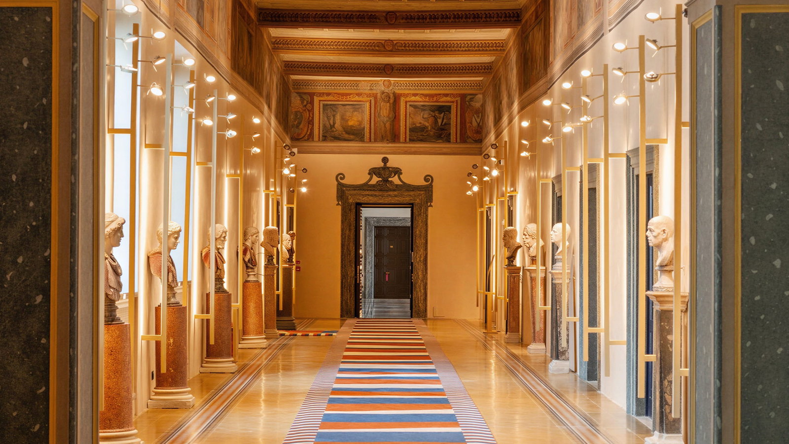 Ornate first floor hallway at Palazzo Talìa with marble busts on columns lining the walls. A colorful striped carpet leads to a distant doorway. Warm lighting and detailed decor.