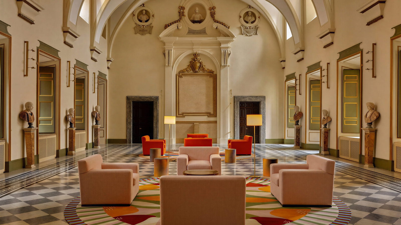 Elegant great hall of Talìa Suite at Palazzo Talìa with arched ceiling, ornate walls, and statues. Colorful armchairs on a patterned rug create a warm, inviting atmosphere.