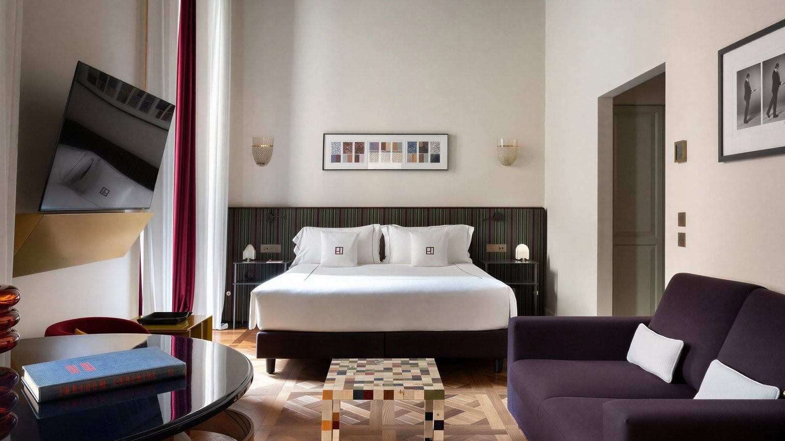 Modern Talia suite bedroom at Palazzo Talìa with a plush double bed, purple sofa, and multicolored cube table. Art on the wall, wooden floor, and a tilted TV. Calm ambiance.