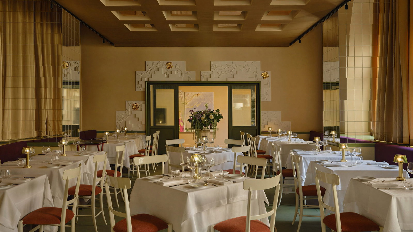 Elegant Tramae restaurant dining room at Palazzo Talìa with white tablecloths, red-cushioned chairs, and soft lighting. Floral centerpiece, warm beige decor, and mirrored walls.