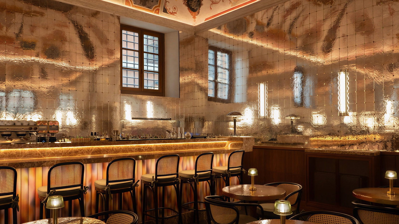Elegant Bar della Musa interior at Palazzo Talìa with reflective metallic walls, ornate ceiling, and warm lighting. Barstools line a lit counter. Cozy ambiance.