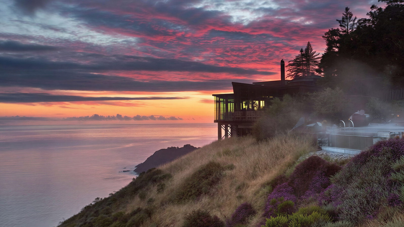 Clifftop exterior of Sierra Mar at Post Ranch Inn overlooking ocean at sunset, with vibrant pink and purple sky. Mist rises near purple flowers and grasses on hillside.