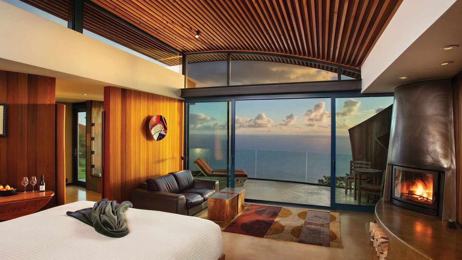 Cliff house bedroom at Post Ranch Inn with ocean view, wooden walls, large windows, fireplace, bed with gray towel, couch, and colorful rug. Cozy and serene setting.