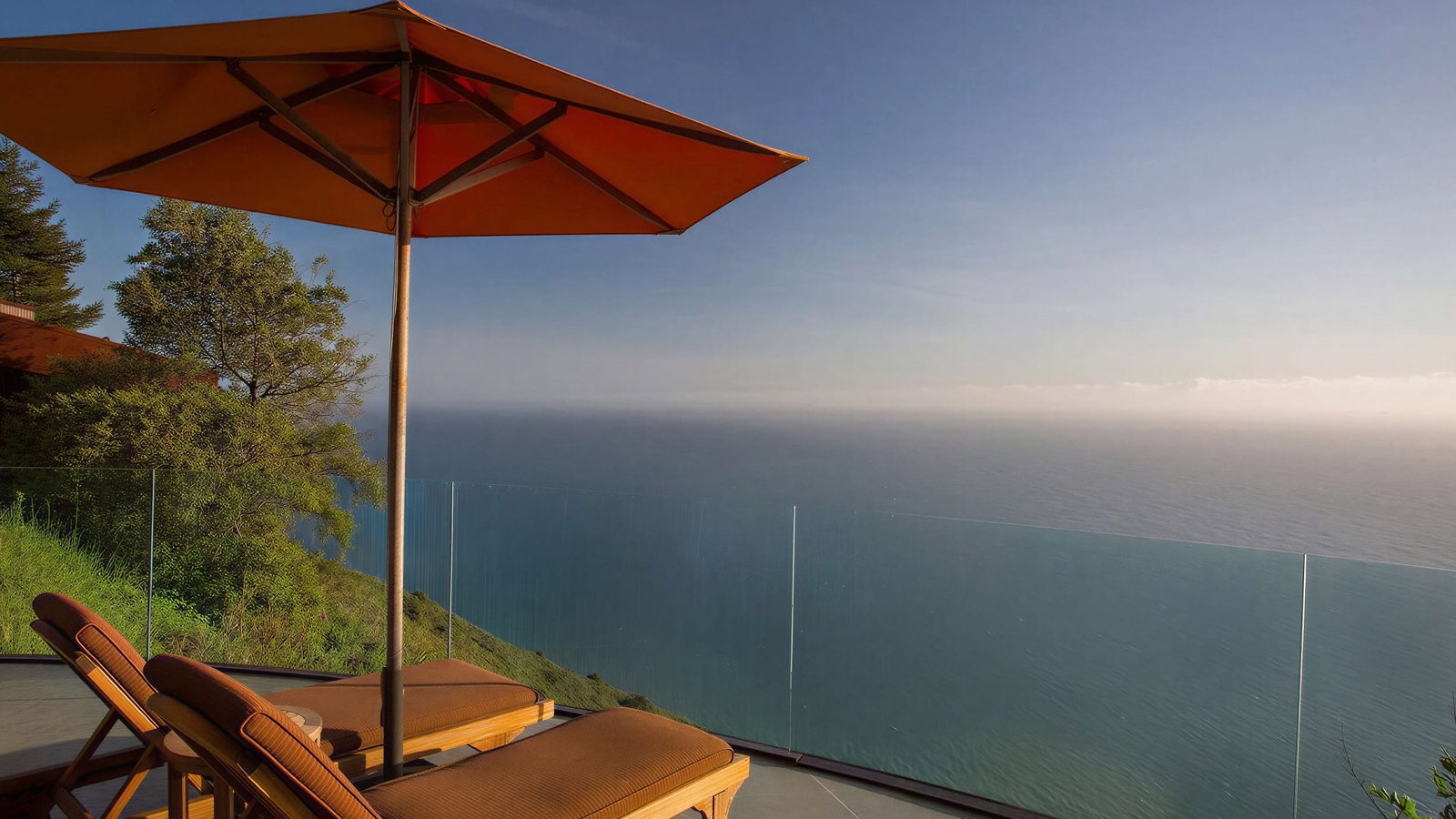 Cliff House deck at Post Ranch Inn. Two lounge chairs under an orange umbrella overlook a calm ocean. Glass railing and green foliage enhance the serene setting.