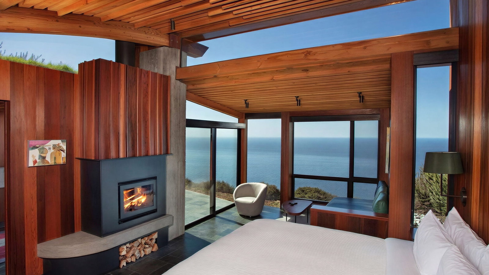Ocean House Northern bedroom at Post Ranch Inn with wood paneling, a lit fireplace, and ocean view through large windows. White bedding, cozy chair, and calm ambiance.