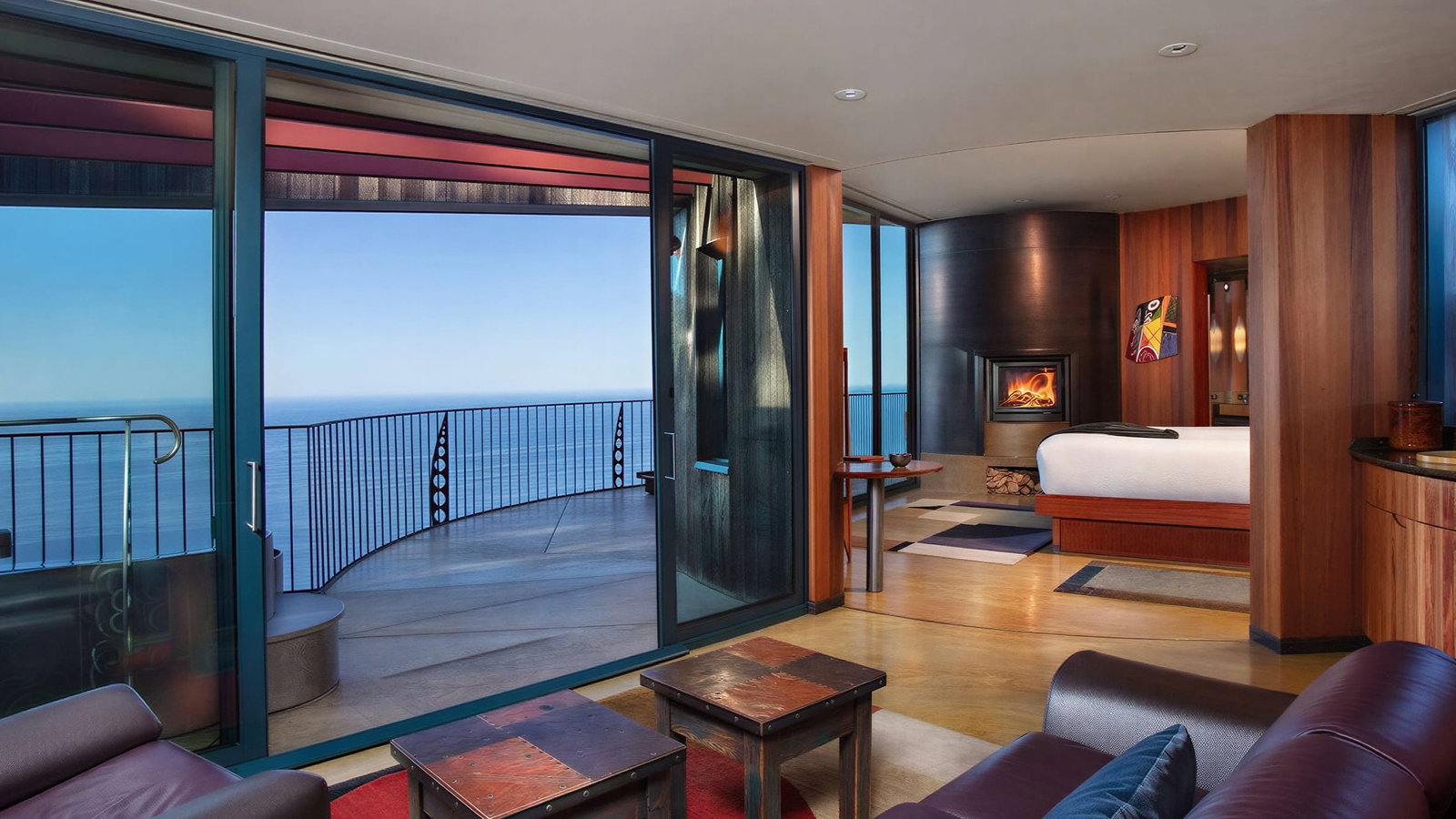 Pacific Suite interior at Post Ranch Inn with ocean view, brown couch, colorful rug, and fireplace. Glass doors open to a balcony under a clear blue sky.
