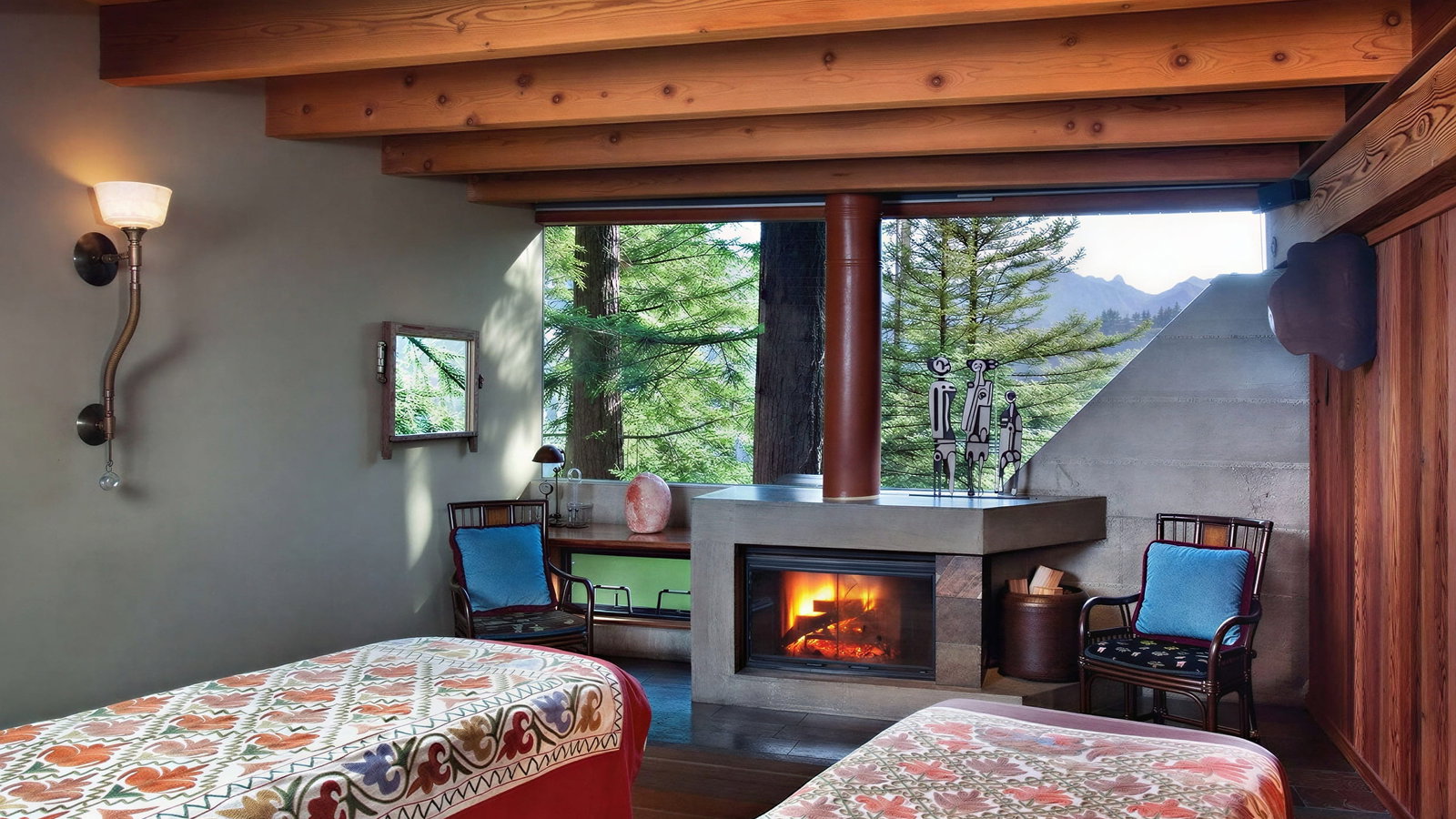 Cozy Post Ranch Inn spa room with two patterned massage beds, a lit fireplace, and a window view of trees. Wooden beams and soft lamp lighting.