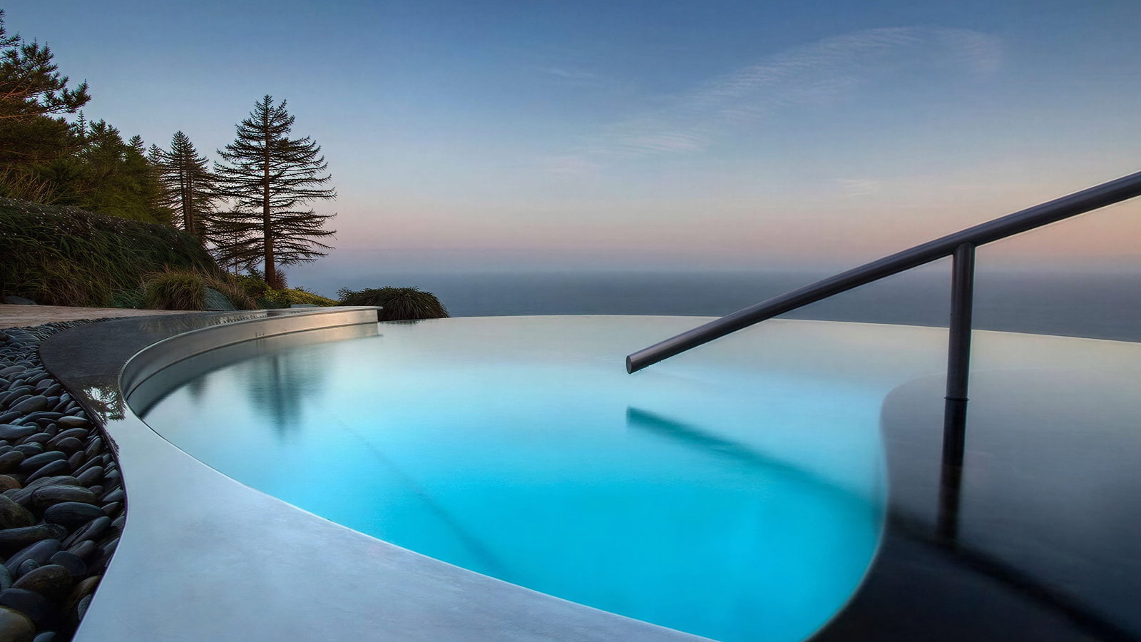 Meditation Infinity pool at Post Ranch Inn overlooking ocean at sunset, surrounded by pebbles and trees. Calm water reflects pink and blue sky, creating a serene mood.