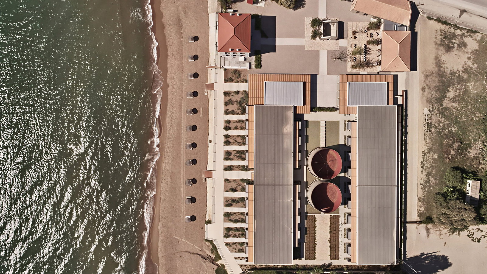 Aerial view of Dexamenes Seaside Hotel with buildings, pathways, and umbrellas along a sandy shore. Waves gently hit the beach.